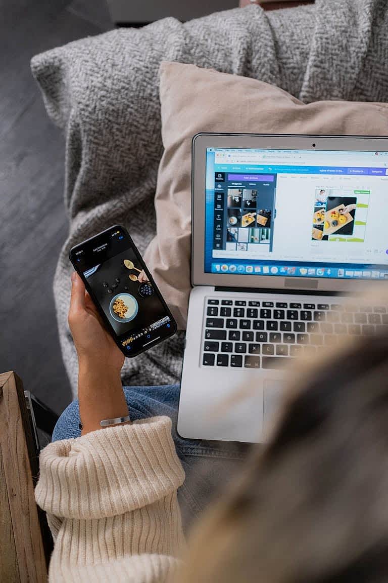 A woman multitasks, using a laptop and smartphone for design work, sitting on a sofa indoors. Bakı web tasarım, seo ajansı