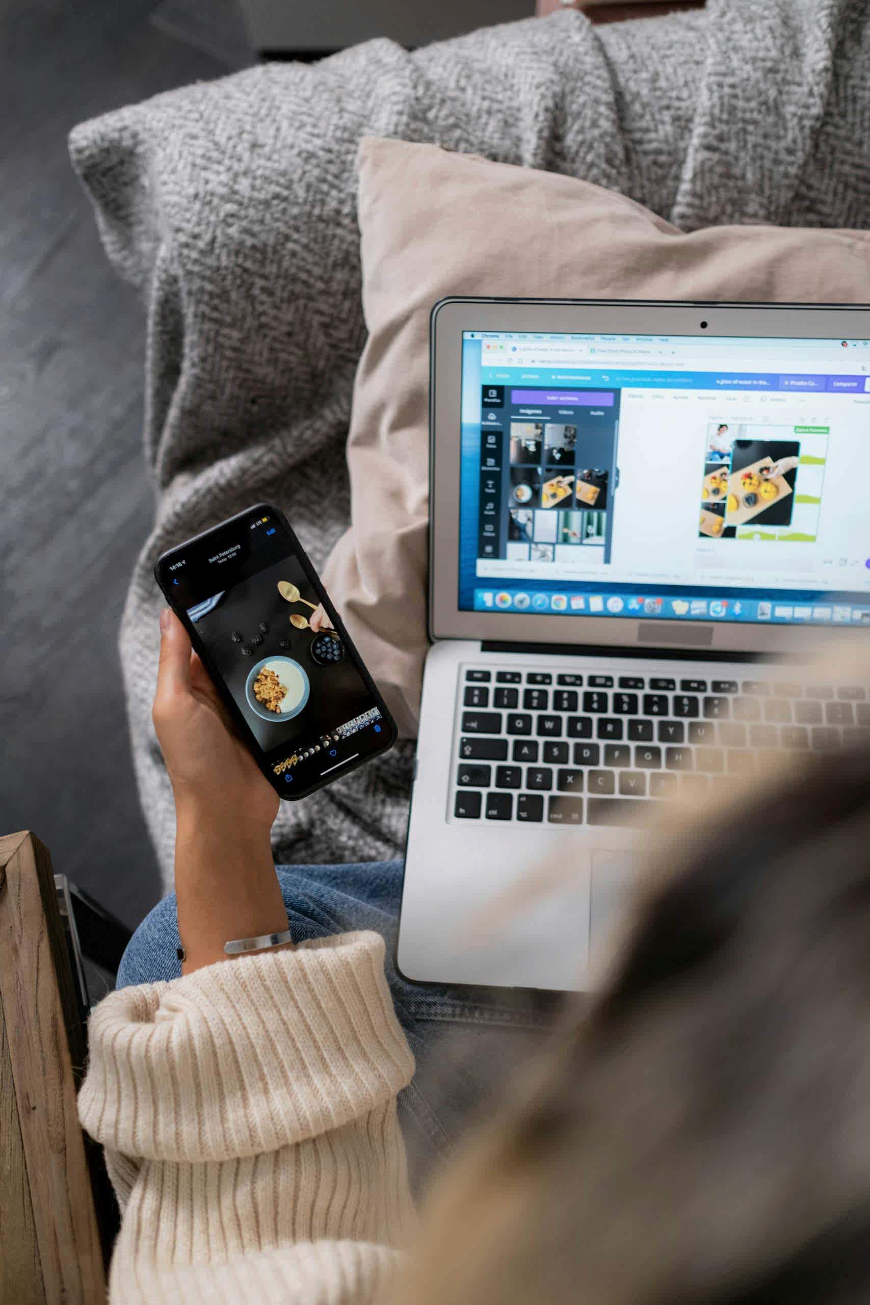 A woman multitasks, using a laptop and smartphone for design work, sitting on a sofa indoors. Bakı web tasarım, seo ajansı
